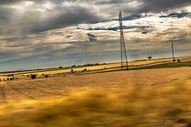 A wide rural landscape shows tall electricity pylons crossing a green field beneath a vivid blue sky. The scene conveys energy infrastructure, openness, and modern contrast with nature.