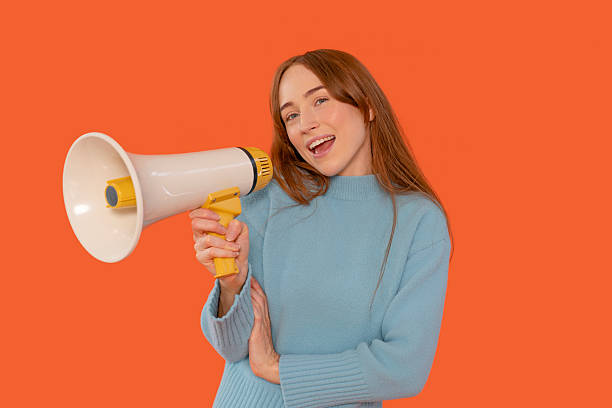 istock Cheerful woman holding megaphone while standing against bright orange background, conveying enthusiasm and energy in vibrant setting 2234482962
