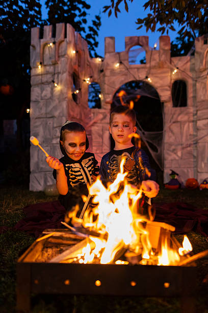 Children dressed in Halloween costumes gather around a campfire at night, roasting marshmallows over the flames. The scene captures the warmth of the firelight, festive spirit, and childhood joy during the spooky holiday celebration.