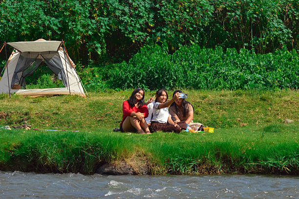 A group of teenage girls sit on grass, smiling for a selfie with their camping tent and lush riverside plants in the background.