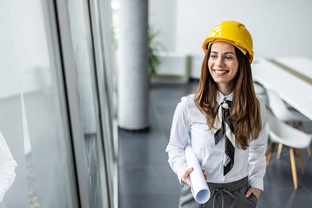 istock Smiling female architect holding blueprints and wearing hard hat in office 2234490010