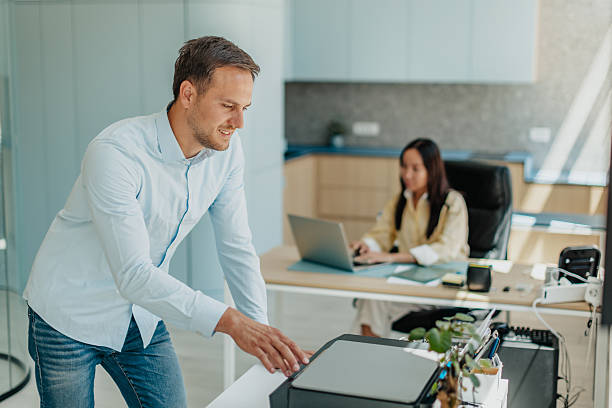 Two professionals engage in their tasks within a contemporary office setting. A man is setting up a printer while a woman works diligently on her laptop, showcasing teamwork and productivity.
