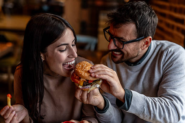 istock Happy couple sharing burger at restaurant table: woman taking bite while man holds it for her 2234494478