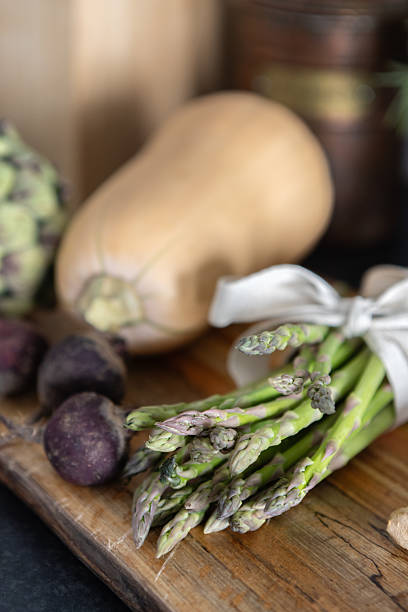 A bundle of fresh green asparagus tied with fabric surrounded by seasonal vegetables, including beets and squash, placed on a rustic wooden cutting board in a close-up kitchen setting.