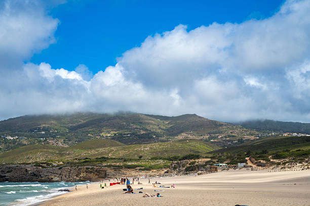 19 August 2025 Praia do Guincho, Portugal. Scenic view of Praia do Guincho in Portugal, with golden sand, beachgoers, and the Serra de Sintra mountains in the background under dramatic clouds. A popular coastal destination for relaxation, surfing, and nature lovers.