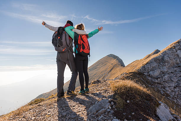 Two hikers embrace at a mountain summit, relishing the spectacular view under a clear blue sky during the day.