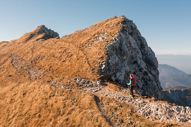 A hiker explores rocky mountain paths amidst golden grasses on a clear autumn day with distant peaks visible.