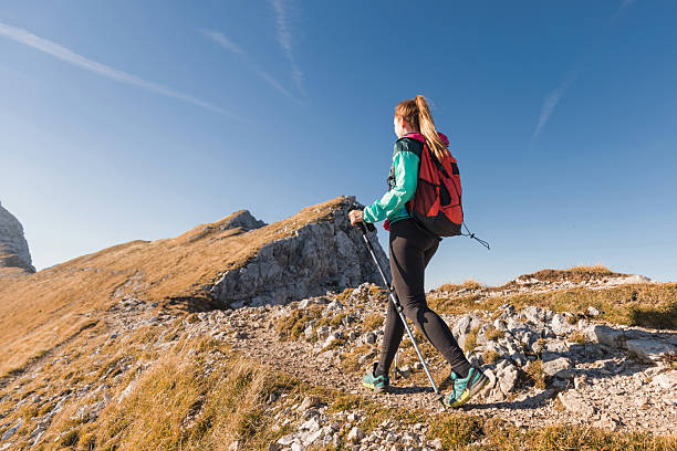 A woman enjoys a sunny hike in the mountains, wearing outdoor gear and navigating a rocky trail.