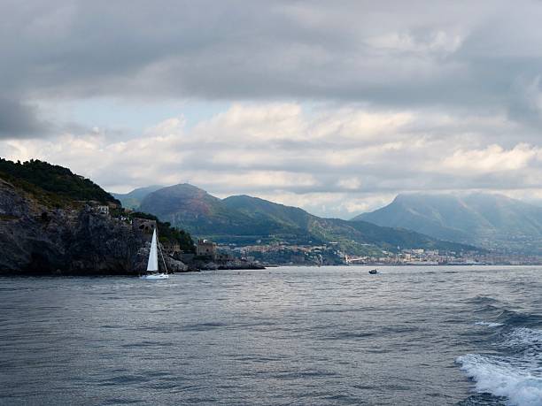 A scenic view of the Amalfi Coast near Salerno, Italy, with dramatic mountains and houses clinging to the hillsides under a cloudy sky. A ferryboat is docked at the harbor, with calm sea in the foreground
