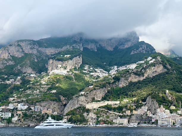 A panoramic view of a coastal town on the Amalfi Coast, with its characteristic white houses and buildings clinging to the lush, green mountainsides. The peaks are partially obscured by dramatic, low-lying clouds. The sea is calm in the foreground.