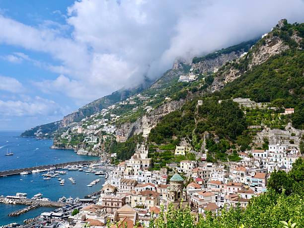 A high-angle, panoramic view of the town of Amalfi on the Amalfi Coast, Italy. The image captures the town's harbor filled with boats, a beach with umbrellas, and the iconic houses built on the steep green mountains