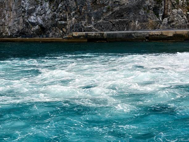 A dynamic close-up of foamy blue and turquoise water near a rocky cliffside. The swirling patterns and white foam of the waves capture the natural power and texture of the sea, creating a powerful abstract maritime scene.