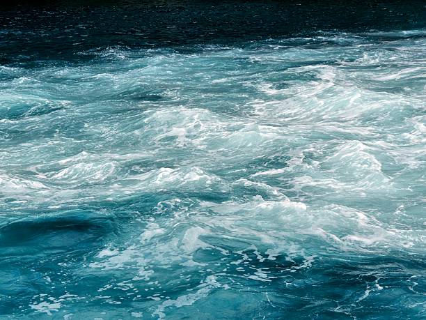 A dynamic close-up of foamy blue and turquoise water near a rocky cliffside. The swirling patterns and white foam of the waves capture the natural power and texture of the sea, creating a powerful abstract maritime scene.