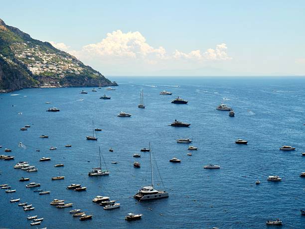 A high-angle, panoramic view of the vast Tyrrhenian Sea, with dozens of boats scattered across the tranquil blue waters under a clear sky. Lush green foliage from the coastline frames the scene, conveying a sense of serene maritime beauty.