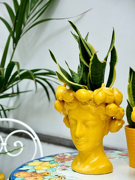 A vibrant yellow ceramic head-shaped planter, adorned with a snake plant, sits on a colorful tiled table outdoors, evoking Mediterranean or coastal decor and gardening