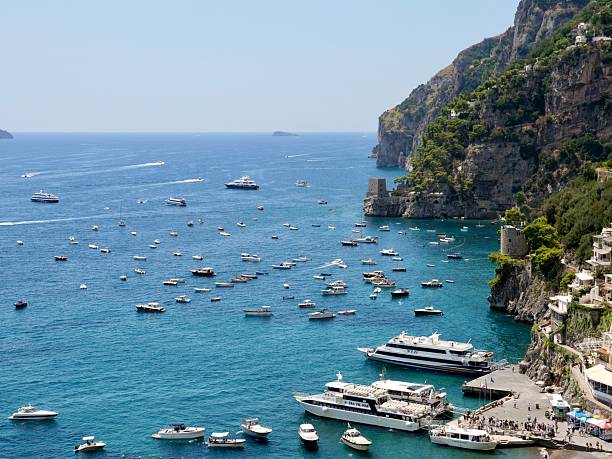 A vibrant panoramic view of Positano, Italy, showcasing its colorful buildings cascading down the steep cliffs to the azure Tyrrhenian Sea. Boats dot the water, and the beach teems with people, embodying a lively summer day on the iconic Amalfi Coast