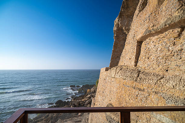 View of a rugged stone wall on the cliffs of Azenhas do Mar, Portugal, with the Atlantic Ocean stretching into the horizon under a bright blue sky.