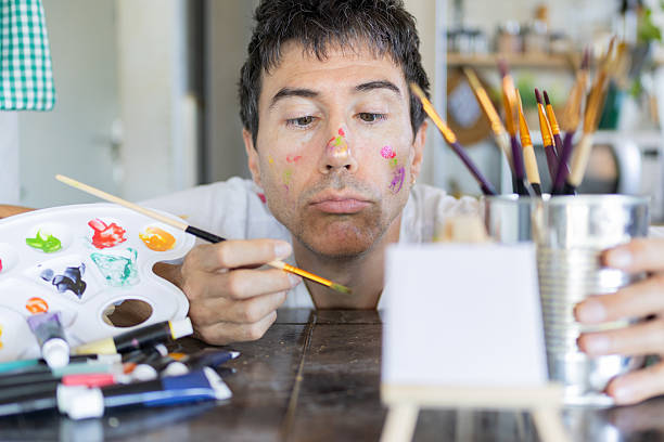 Mid adult male artist concentrating while painting a small canvas on a wooden table, surrounded by colorful paints and brushes in his home studio