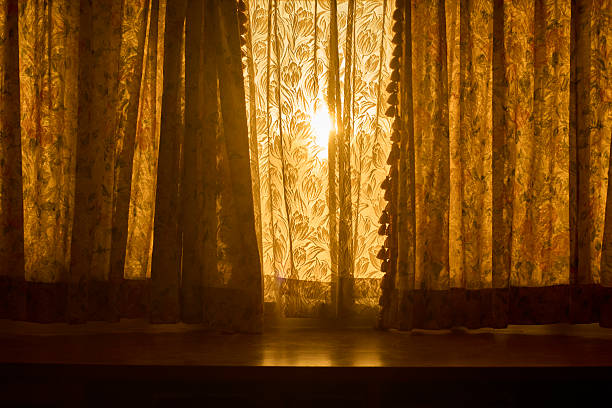 Horizontal interior scene of vintage floral curtains with a decorative tassel trim, photographed backlit as warm sunlight glows through the patterned fabric. The folds and pleats create texture and a nostalgic, cozy atmosphere suitable for themes of home, privacy, hospitality, and morning calm. Soft golden tones provide ample copy space for titles or branding. Works for editorial features, lifestyle and décor content, brochures, ads, and website hero images. No people and no visible trademarks; full frame.