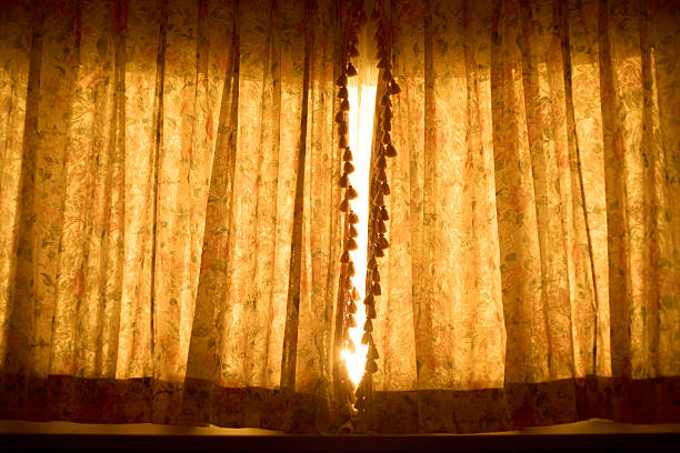Horizontal interior scene of vintage floral curtains with a decorative tassel trim, photographed backlit as warm sunlight glows through the patterned fabric. The folds and pleats create texture and a nostalgic, cozy atmosphere suitable for themes of home, privacy, hospitality, and morning calm. Soft golden tones provide ample copy space for titles or branding. Works for editorial features, lifestyle and décor content, brochures, ads, and website hero images. No people and no visible trademarks; full frame.