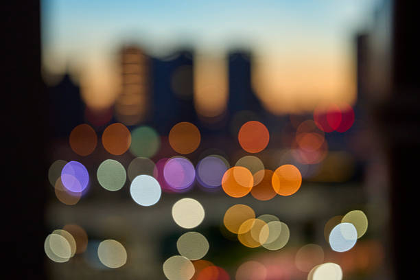 Vertical, abstract cityscape photographed at dusk/twilight with the skyline rendered out of focus. Large, multicolored bokeh circles—from street and building lights—float against a soft sunset gradient, shifting from warm oranges to cool blues. The image conveys nightlife, commute, mood, creativity, and urban energy while leaving generous copy space for headlines or logos. Ideal as a background for editorials, websites, ads, presentations, and social posts. No people and no visible trademarks; portrait orientation; full frame.