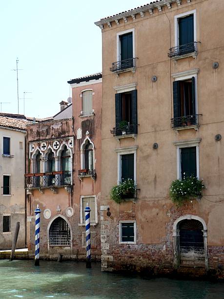 Historic, weathered buildings directly on a small Venetian canal. Features traditional Gothic windows, colorful plaster, and striped mooring poles (pali) under a bright sky
