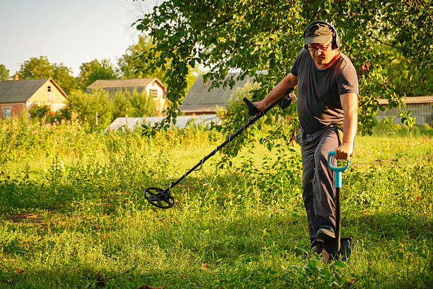 istock A man uses a metal detector in a vibrant green field surrounded by trees and rustic structures. The summer sun casts a warm glow as he carefully scans the ground, focused on finding hidden treasures 2234530180