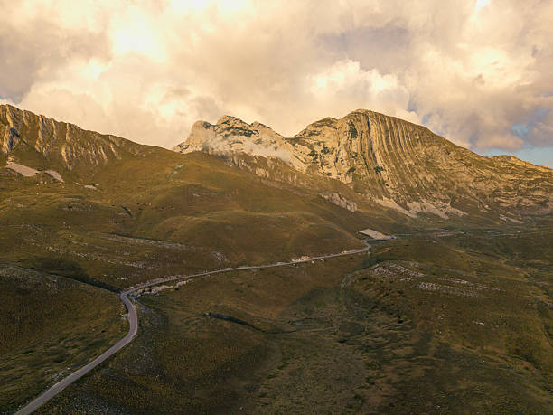 Aerial view of rugged mountain terrain with a winding road cutting through grassy slopes. Clouds hover near rocky peaks under soft sunlight. Scenic natural landscape with open space.
