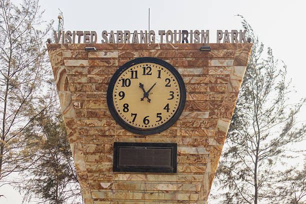 Golden textured clock monument standing tall under sunlight with coastal trees and seashore in background.