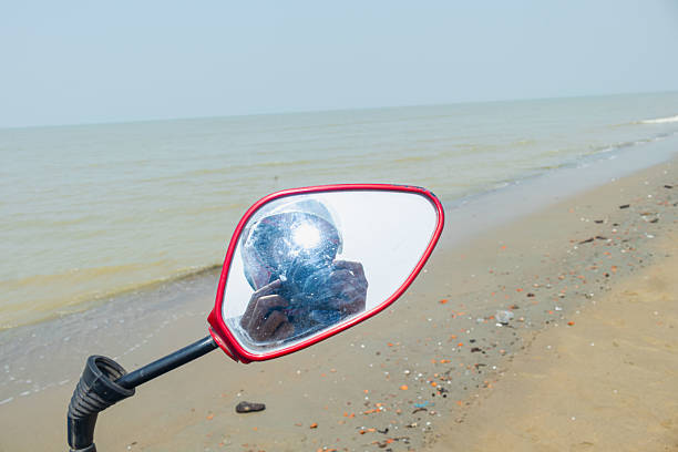 Unique red motorcycle mirror captures photographer’s reflection with serene ocean waves in the background, blending vibrant foreground detail with tranquil coastal scenery.