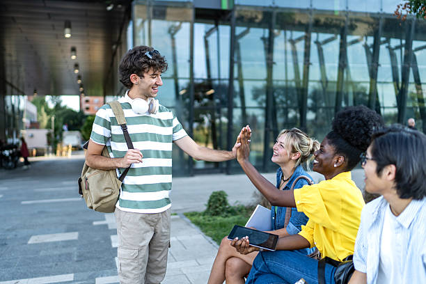 Cheerful young adults sitting on a bench in front of their college, greeting each other with high fives, celebrating friendship and connection in a vibrant campus atmosphere