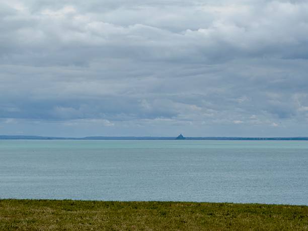 Horizontal landscape photo of the tiny Mont St Michel across the calm blue sea on the distant horizon as seen from a grassy hilltop in Cancale, Brittany, France.