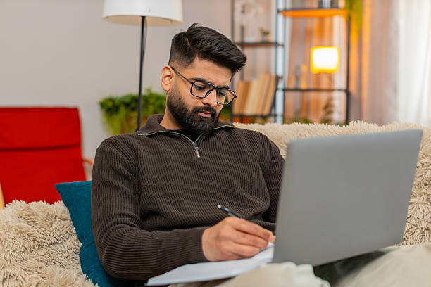 istock Indian man writing notes from laptop into notebook at home on sofa with focused look studying online 2234571120