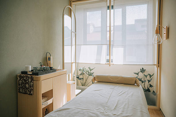 serene interior of a spa room featuring a single massage bed, soft daylight through sheer curtains, and minimalist decor that conveys peace and relaxation. Location: wellness center.