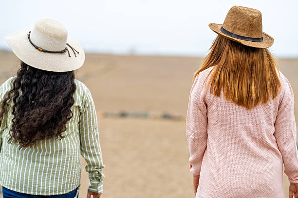 Back view of two beautiful young women friends standing together in autumn park, stylish girlfriends in hat and sweater outdoors looking at trees with yellow leaves, nature background