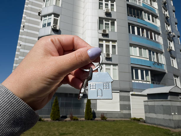 istock A woman's hand holds a house key in plain view in front of a tall, elegant modern building rising in the background. 2234604443