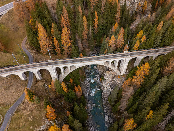 A stone arch railway bridge crosses a deep valley surrounded by autumn-colored pine and larch trees. Scenic mountain landscape with winding roads and cloudy sky. Mountain travel and nature.