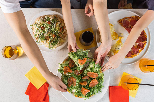 Group of diverse hands reaching for fresh salad and pizza on a table, with colorful napkins and drinks, creating a lively dining atmosphere and shared meal experience