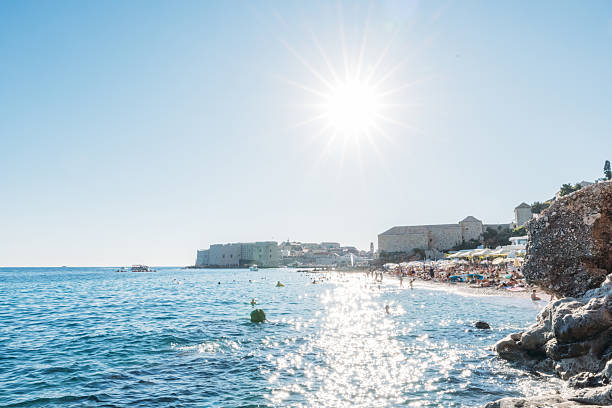 Bright sunny day at a crowded beach with sparkling blue water and rocky shoreline. People relaxing and swimming near a historic coastal town with old stone buildings. Summer vacation scene.