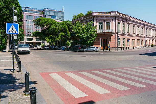 A lively urban intersection features a crosswalk and a mix of vehicles parked along the street. Modern and classic buildings line the area under a clear blue sky.