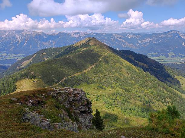 An expansive, high-altitude view showcasing a hiking trail that follows the long, grassy crest of an alpine ridge. The foreground features a rugged rock outcropping and heather-covered slopes. The distant background reveals multiple layers of majestic mountains and the wide, cultivated valley floor below (the Enns Valley). The scene is set under a dramatic sky with scattered white clouds, characteristic of the beautiful Schladming-Dachstein region in Austria.