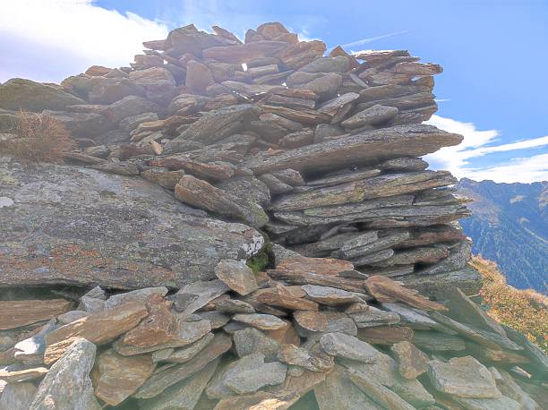 A close-up of a carefully stacked stone cairn on a mountain ridge, highlighted by rugged, weathered rocks and a bright blue sky. The photo captures the cairn in its natural alpine environment, with misty peaks and lush valleys visible in the background. This image symbolizes trekking, navigation, and the character of wild mountainous landscapes, ideal for adventure, travel, or outdoor-themed content.