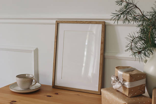 istock Christmas Scandinavian interior. Winter artistic composition. Blank vertical wooden picture frame mockup, cup of coffee on table. Pine tree branches in vase. Blurred gift boxes. White wall background 2234629421
