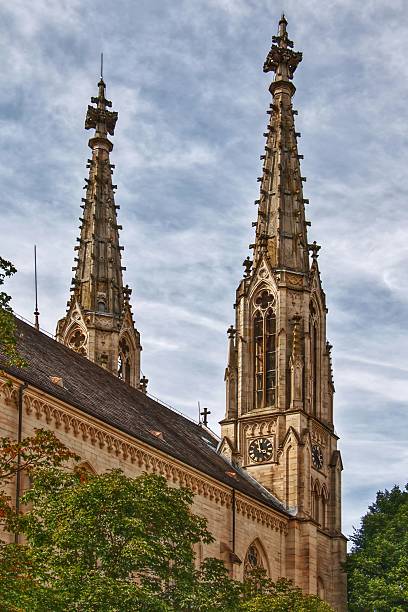 A majestic Gothic-style cathedral with twin ornate spires rises above the main square of Baden-Baden, Germany. Surrounded by lush trees and set against a dramatic cloudy sky, it stands as a historic and architectural landmark.