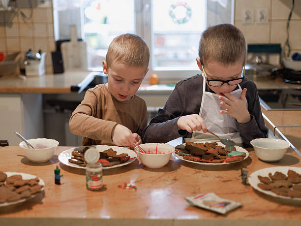 Two children decorate gingerbread cookies with icing, one tasting the glaze while the other focuses on decorating. Fun family Christmas tradition in the kitchen.