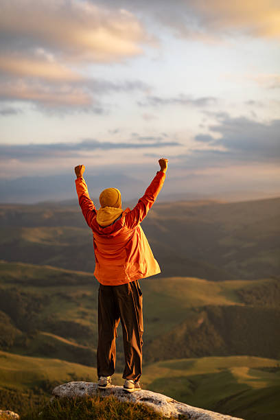Mid adult man stands on mountain summit with arms raised, looking at sunset and celebrates success