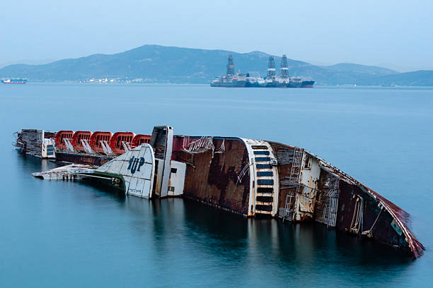 Rusty shipwreck in Elefsina bay, Attica, Greece, half-sunken in calm sea waters, with offshore oil platforms and tankers in the industrial background.