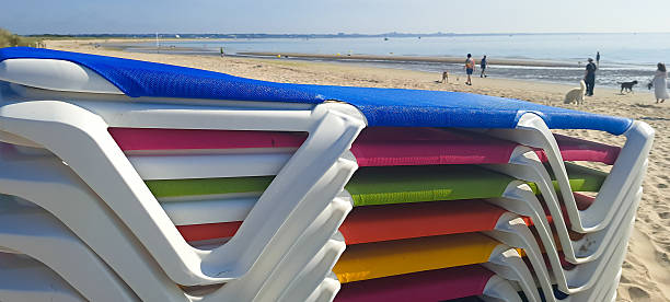 Stack of brightly coloured sun loungers ready for hire on the beach on a sunny summers day. Holiday makers hire the seats for the day to allow them to sun bath and get a sun tan whilst relaxing on vacation.