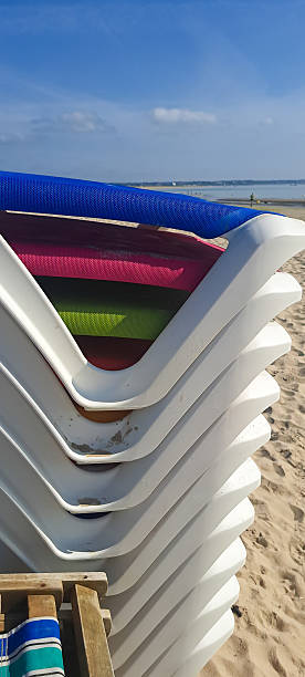Stack of brightly coloured sun loungers ready for hire on the beach on a sunny summers day. Holiday makers hire the seats for the day to allow them to sun bath and get a sun tan whilst relaxing on vacation.