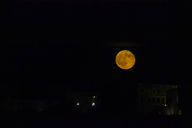 The brilliance of an orange moon illuminates a night, set against an urban skyline. A calm yet mysterious ambiance transcends as the silent metropole outlines enhance the natural beauty of the scene.